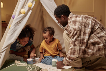 Caring African American father treating little kids to hot chocolate drinks with marshmallows on top while children having fun inside teepee tent in bedroom © Seventyfour