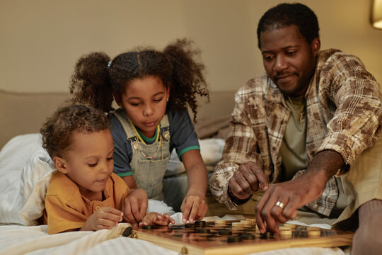 Medium shot of two African American kids moving pieces on board while playing checkers game with dad lying in bed at home, happy family scene