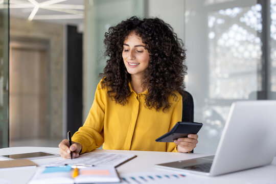 Successful satisfied female financier doing paperwork inside office. Businesswoman holding calculator in hands and smiling writing down filling out tax forms and investment reports. - Powered by Adobe