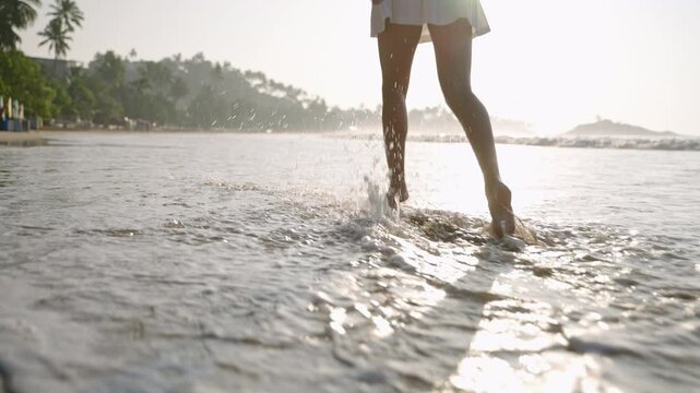 Female feet walking barefoot in sea surf waves. Person's legs running along sandy coast. Attractive woman in mini skirt strolling and enjoying sunset at the seaside on vacation. Low angle.