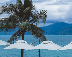 Palm trees and white umbrellas frame a picturesque view of the turquoise ocean and distant mountains under a partly cloudy sky