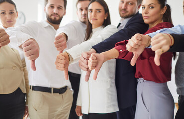 Group of serious professionals standing together in office, all giving thumbs down gesture. Team of young business people expressing dissatisfaction or disapproval, showing dislike and rejection.