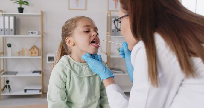 Child pediatrician doctor examining little girl throat during visit in clinic, holding wooden spatula tongue reviewing patient tonsils, provide professional medical help, otolaryngology pediatric