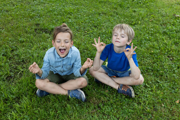 Two cheerful boys are having fun sitting on grass, imagining themselves at yoga classes as adults. Active recreation in summer in the park or in the school yard. positive, joy of childhood, pampering
