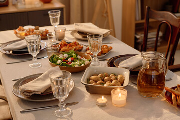 High angle shot of fresh Greek salad and baked baby potatoes with olive oil mixed with herbes de Provence surrounded by clean and polished tableware, lit wax candles and snack on table