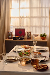 Vertical shot of table covered with white tablecloth and served with homemade food, table located near wooden dresser with speakers and retro vinyl record