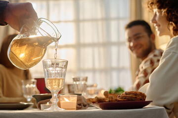 Low angle shot of unrecognizable male hand holding crystal glass lit by sunray, he pouring out glass of apple juice while his guest sitting at festive table and chatting