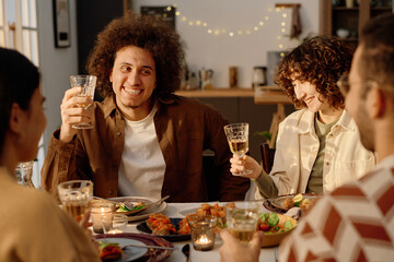 Curly haired young adult biracial man holding glass with apple juice while smiling widely during funny and warm conversation with his friends