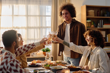 Wide shot of biracial friends clinking glasses with champagne above served festive table with homemade delicious food and tasty appetizers
