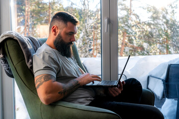 Concentrated young man with beard and tattoos using laptop while sitting in comfortable armchair...