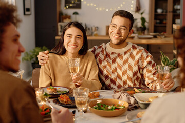 Young adult Caucasian couple holding glasses with champagne hugging and looking at their friends while celebrating house-warming