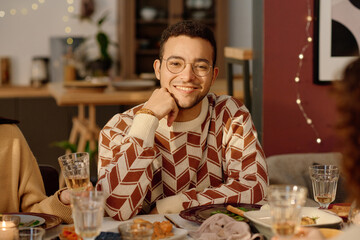 Medium close up portrait of young adult biracial man dressed in sweater with ornaments leaning his head on hand and smiling while looking at camera