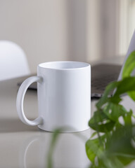 White coffee mug on grey office table against laptop, glasses and plant closeup, mockup