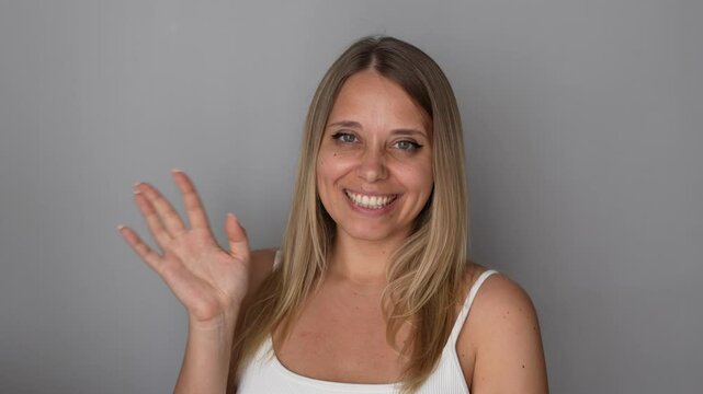Young happy smiling caucasian blonde woman waving her hand in greeting saying "Hi" on a grey background. Hello gesture