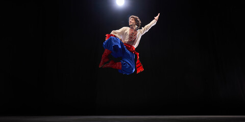 Male dancer in traditional embroidered shirt, blue trousers, and red boots mid-air leap, showcasing cultural heritage and energy against black studio background. Concept of folk style, art and history