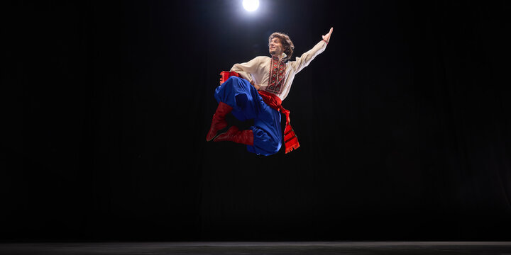 Male dancer in traditional embroidered shirt, blue trousers, and red boots mid-air leap, showcasing cultural heritage and energy against black studio background. Concept of folk style, art and history