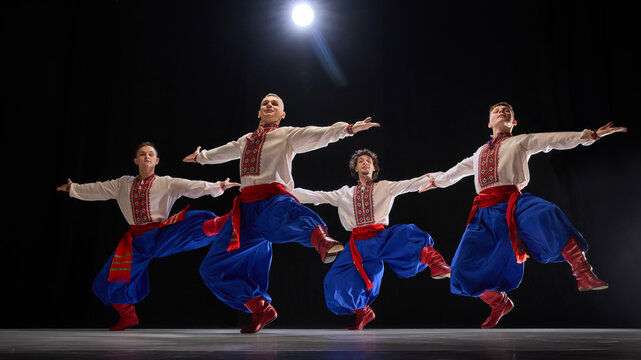 Four male dancers in formation, arms outstretched, dressed in embroidered shirts, blue trousers, and red boots, highlighting unity and tradition against black background. Concept of art and history. - Powered by Adobe
