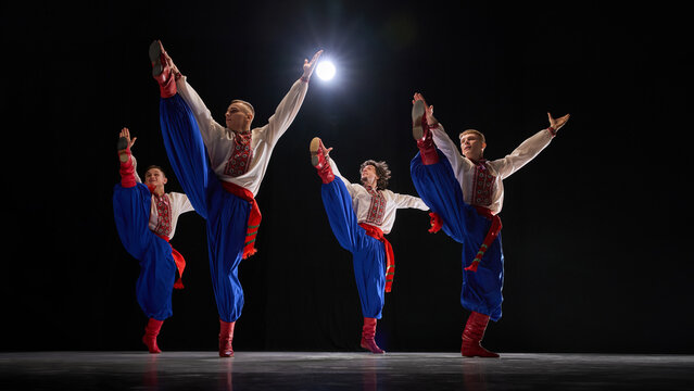 Male dancers in powerful high kick pose, wearing embroidered shirts, blue trousers, and red boots, showcasing tradition and precision against black studio background. Concept of art, history.