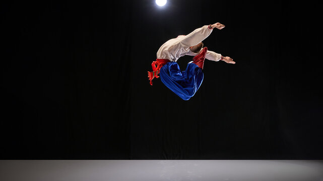 Male dancer executing backward jump, dressed in traditional embroidered shirt, blue trousers, and red boots against black studio background. Concept of traditions, folk style, people, art and history.