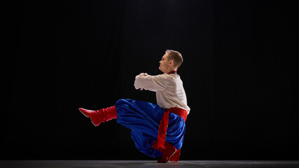 Male dancer in deep squat with one leg extended, arms elegantly balanced, showcasing agility and tradition against black studio background. Concept of traditions, folk style, people, art and history.