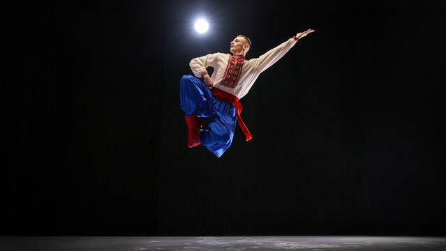 Male dancer in controlled high jump, body extended in confident pose, traditional attire flowing to symbolize cultural pride against black background. Concept of traditions, folk style, art. Ad