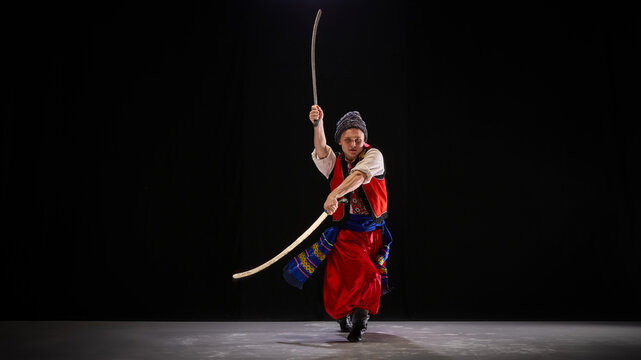 Dancer in Ukrainian folk outfit, confidently wielding dual swords in traditional Hopak stance, embodying historical spirit on dark background. Concept of traditions, art and history. Ad