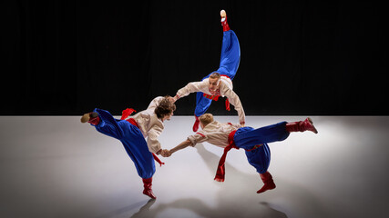 Three dancers hold hands mid-performance, creating motion with high kicks and synchronized movements, symbolizing unity in Hopak. Concept of traditions, folk style, art and history. Ad