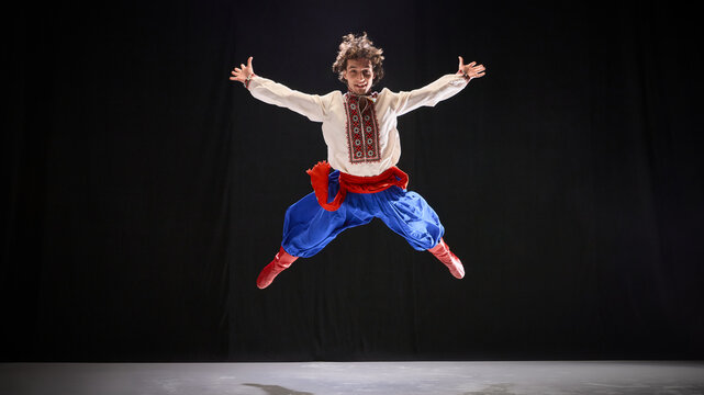 Dancer in embroidered shirt and blue Ukrainian folk attire, jumping wide legs and hands against black background, emphasizing athleticism of Ukrainian Hopak. Concept of traditions, folk style. Ad