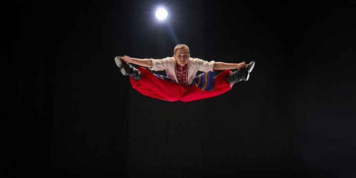ale dancer in bright red trousers performs airborne split jump, arms outstretched, showcasing explosive athleticism of Hopak, against black studio background. Concept of traditions, folk style. Ad