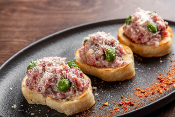 Delicious appetizer featuring three toasted bread slices topped with savory minced meat, cheese, and green peppers, garnished with a sprinkle of spices on a stylish black plate.