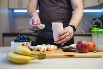 Man preparing a protein drink in a kitchen, holding a portion of whey protein powder in a measuring spoon and a glass of milkshake or smoothie in his hand