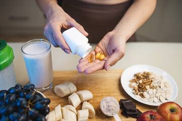 Athletic woman in sportswear is holding omega-3 capsules, fish oil supplements in her hand, protein powder in a measuring spoon, smoothie drink in a glass, banana and grapes on a table in a kitchen
