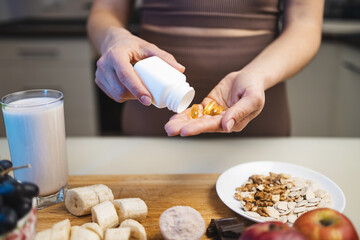 Athletic woman in sportswear is holding omega-3 capsules, fish oil supplements in her hand, protein powder in a measuring spoon, smoothie drink in a glass, banana and grapes on a table in a kitchen