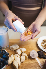 Athletic woman in sportswear is holding omega-3 capsules, fish oil supplements in her hand, protein powder in a measuring spoon, smoothie drink in a glass, banana and grapes on a table in a kitchen