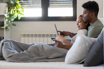 A young couple sits together on a bed, holding a credit card and tablet, smiling as they engage in...