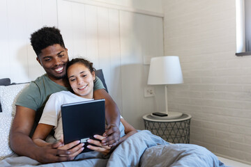 A cheerful couple cuddled together in bed, using a tablet to browse or watch content, enjoying a cozy and relaxing moment in a bright, modern bedroom