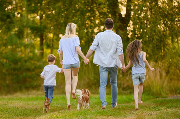 Fototapeta premium Family enjoying beautiful day outdoors, holding hands, running barefoot together on grass, view from back. Family walking happy dogs. Concept of summer weekends, country vacation, being with nature.