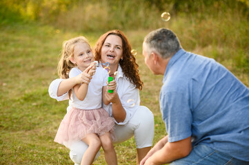 Fototapeta premium Mom and sweet girl blow soap bubbles, sitting on grass in summer, dad is happy to play with them, everyone is laughing and smiling. Concept of summer holidays, carefree, childhood. Children's Day