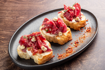 Delicious crostini topped with savory sliced meat, creamy sauce, and fresh herbs, served on a stylish black platter. Perfect for gourmet appetizers or party snacks.