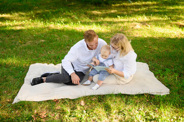 Mom and dad read book to cute child, look at educational pictures in book, sit in park during picnic, on weekend. Loving parents spend time with charming baby, learn, communicate.