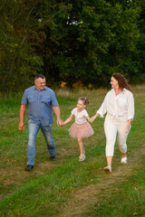 Fototapeta premium Family walking together in summer. Mom, dad, daughter walking in meadow holding hands, smiling happily, girl jumping happily. Family, love and concept of parenthood, happy childhood.