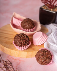 brigadeiro em forminhas de papel rosa sob uma tábua de madeira em um fundo rosa com um pano branco ao lado e um mini vaso atrás


