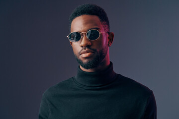 Obraz premium Stylish young man with sunglasses in a turtleneck sweater, expressing confidence against a dark gradient background, ideal for fashion and lifestyle themes