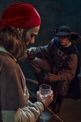 Focus on man, pirate in red bandana standing with thoughtful face with glass of rum, with his mate sitting with a drink in the background in dimly-lit room. Concept of history, adventure, lifestyle