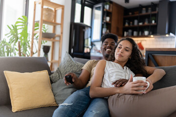 A cheerful couple relaxing on a cozy sofa at home, watching TV together; the man holds the remote while the woman sips a hot drink in a modern, well-lit living room
