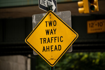 Yellow road sign indicating two-way traffic ahead in an urban area surrounded by buildings and traffic signals during the day