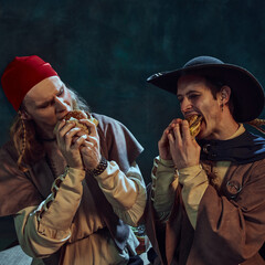 Close-up portrait of tow men, medieval pirates in hat and bandana, passionately eating burgers...