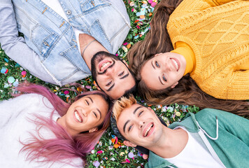 Smiling young people taking selfie lying on the grass at park among the confetti - Girls and boy celebrating and laughing out loud outdoor - Friendship concept with young students having fun together