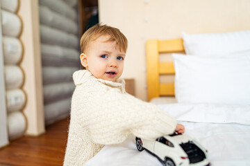 Fototapeta premium Child plays with toy car in cozy bedroom. A young child in a knit sweater engages with a toy car on a bed in a warm, inviting bedroom environment.