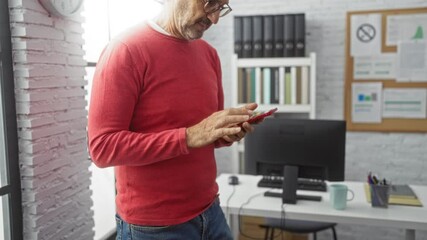 Senior man in office wearing glasses and using smartphone with focused expression stands next to a desk with a computer and documents, suggesting a modern workplace environment. - Powered by Adobe